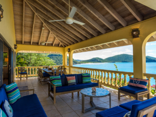 Covered patio with cushioned outdoor seating, a formal dining table and ceiling fans and Mahoe Bay in the background.
