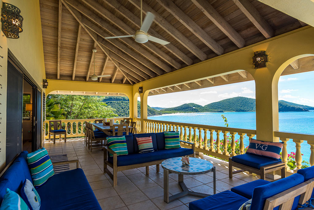 Covered patio with cushioned outdoor seating, a formal dining table and ceiling fans and Mahoe Bay in the background.