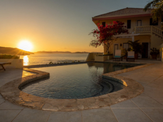 Swimming pool and stone patio outside A Dream Come True Villa with the sun setting on Mahoe Bay in the background.