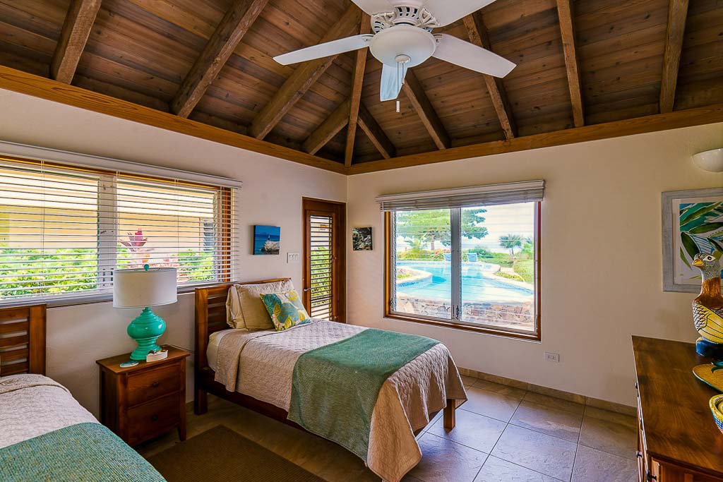 Beachcomber Villa double bedroom with a wood ceiling with ceiling fan and windows looking out on a pool and Mahoe Bay.