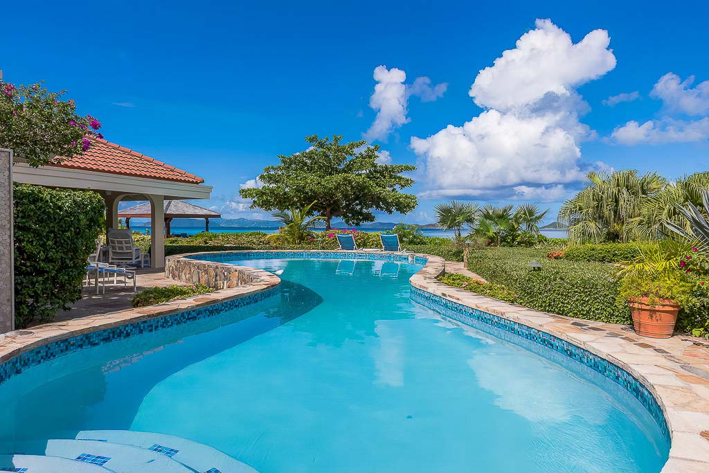 Large, free-form pool at Beachcomber Villa surrounded by shrubs and trees with Mahoe Bay in the background on a sunny day.
