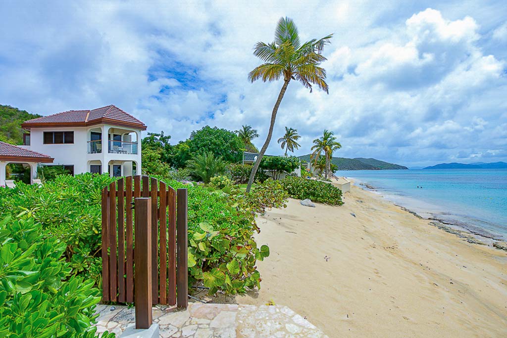 Wood gate on a stone path next to the sandy beach and blue water of Mahoe Bay with palm trees and hills in the background.