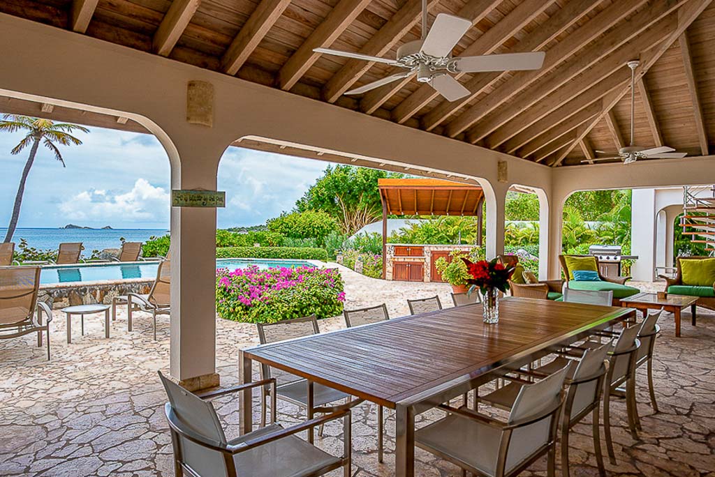 Formal outdoor dining table on a natural stone patio covered by a wood-beam roof with a pool and Mahoe Bay in the background.