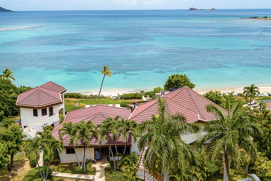 Caribbean Wind Villa surrounded by palm trees in front of Mahoe Bay beach with the crystal blue sea in the background.