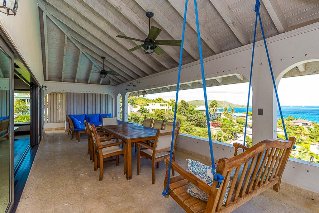 A covered porch with a wooden dining table, a swing seat and ceiling fans with Leverick Bay, Virgin Gorda in the background.