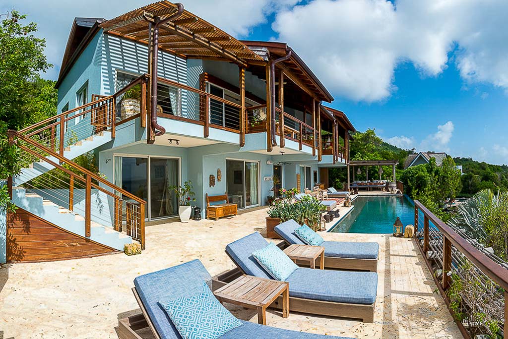 A natural stone patio with lounges and a pool in front of La Vida Villa with multiple glass doors and second level balconies.