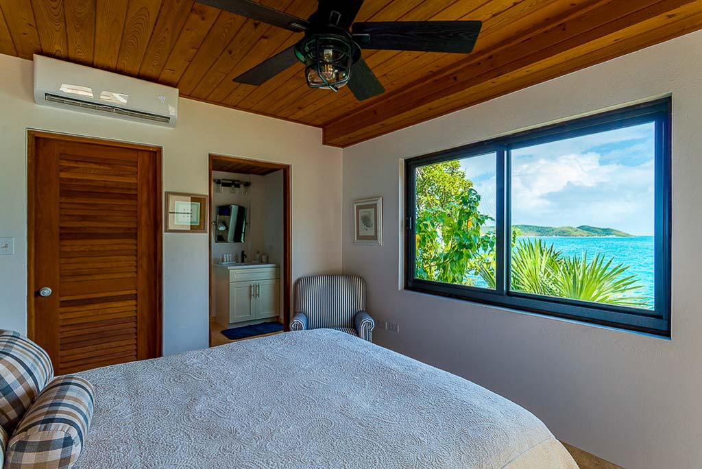Guest bedroom at Rainbow’s End Villa with en suite bathroom, wood ceiling and window looking out on tropical Leverick Bay.