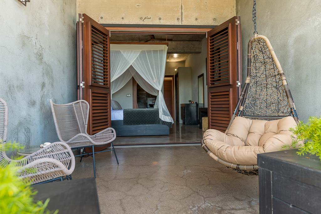 Private patio with a hanging wicker nest chair off a Segura Villa guest room with a king-size bed in the background.
