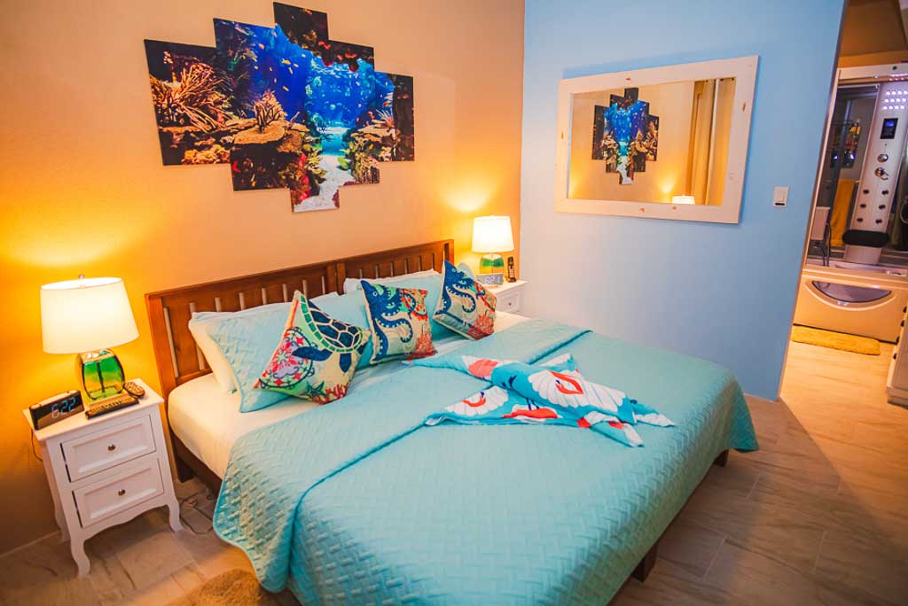 Image 8: 1 Paradise Lane Villa guest room with a king bed, bright tropical art on the wall and a side table with a lamp.