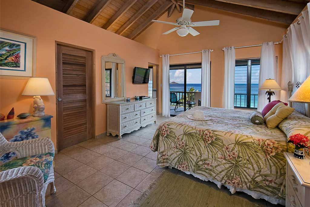 Second master suite with a king bed, bathroom, white bureau and a door leading to a veranda with Mahoe Bay in the background.