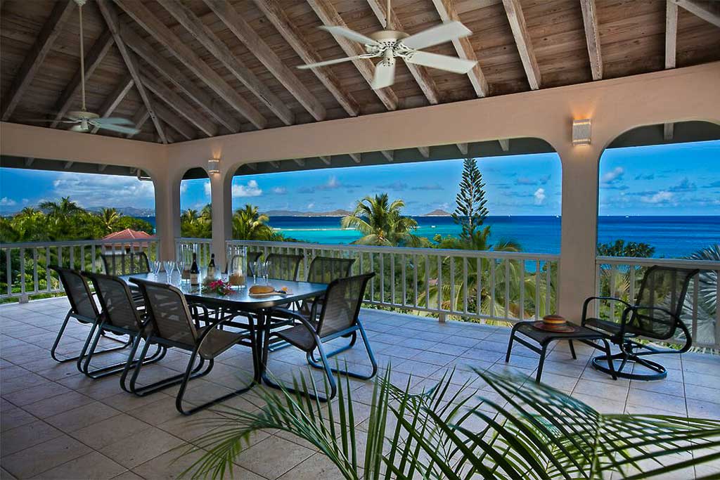 Outdoor dining area on the covered ocean-side porch at Adagio Villa with ceiling fans and a backdrop of sunny Mahoe Bay.