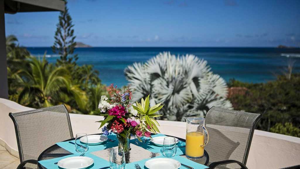Round table set for breakfast on the Adagio Villa patio with palm trees and the blue waters of Mahoe Bay in the background.