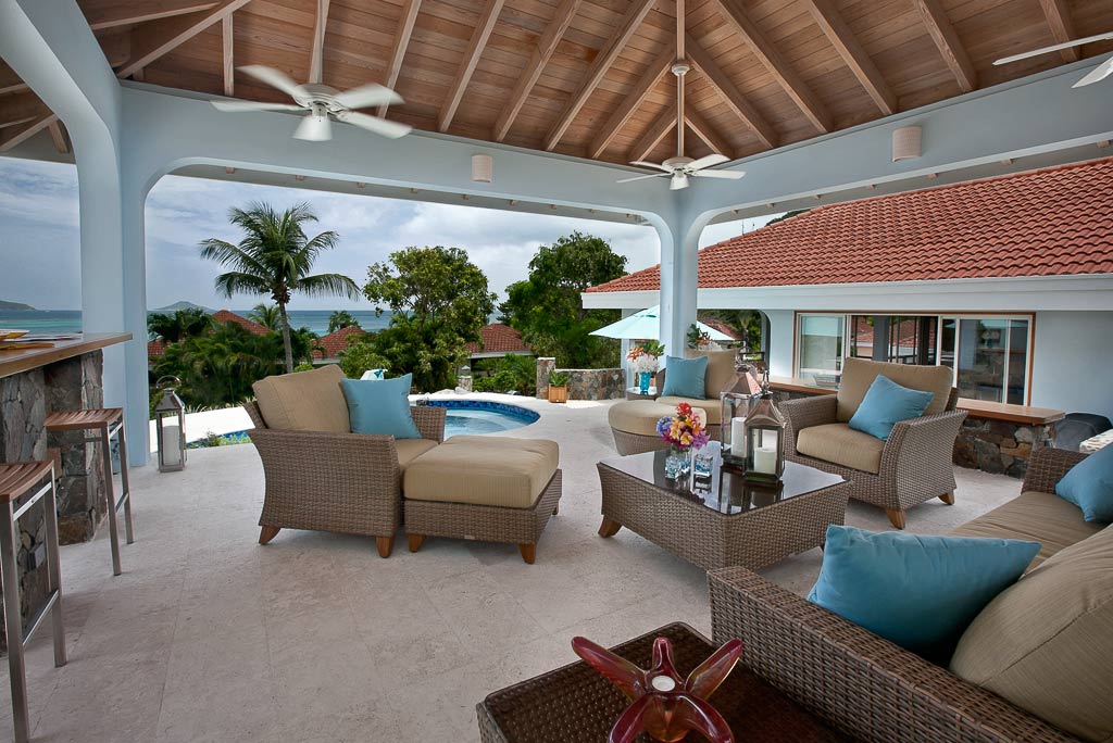 Covered stone patio with a couch and comfortable seating and a wood-beam ceiling with trees and the sea in the background.