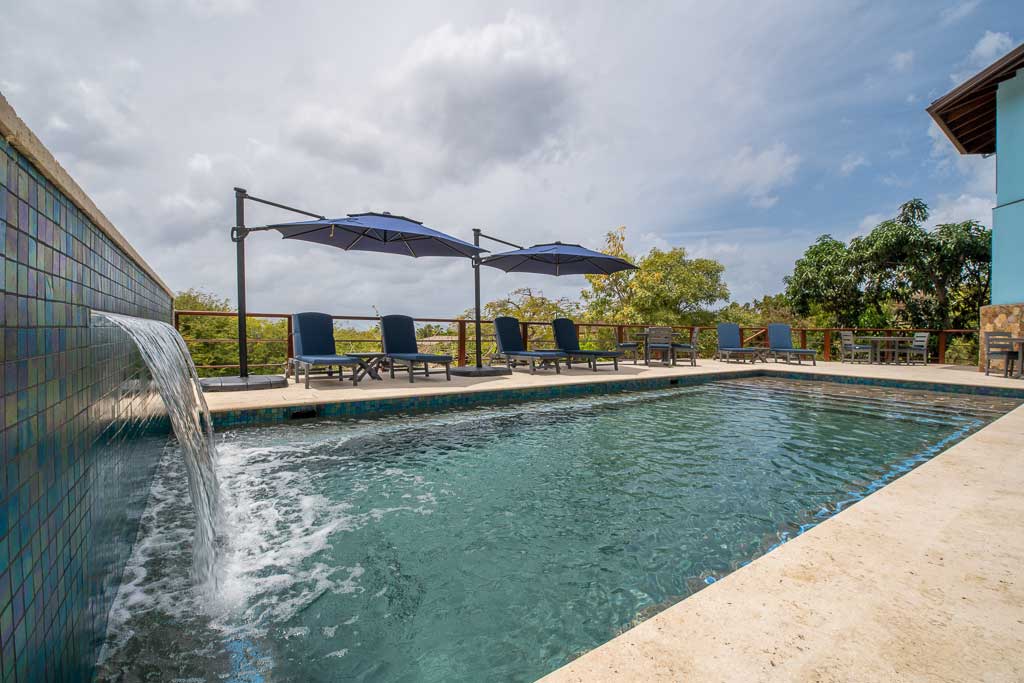 Eden Waters Villa’s swimming pool with waterfall surrounded by a natural stone patio with loungers and umbrellas.