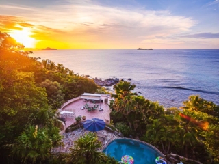 Pool and oceanfront patio at Sundowner Villa surrounded by palm trees with the sun setting on Nail Bay in the background.