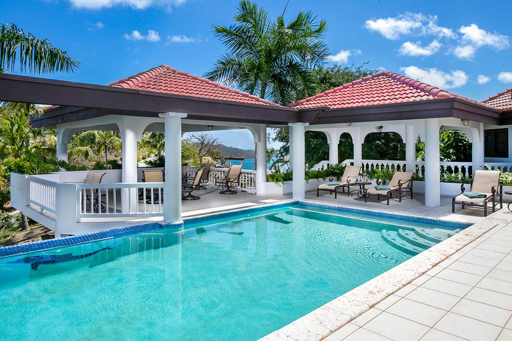 Villa Tamar’s swimming pool surrounded by a white tile sun deck with covered sitting and lounging areas and palm trees.