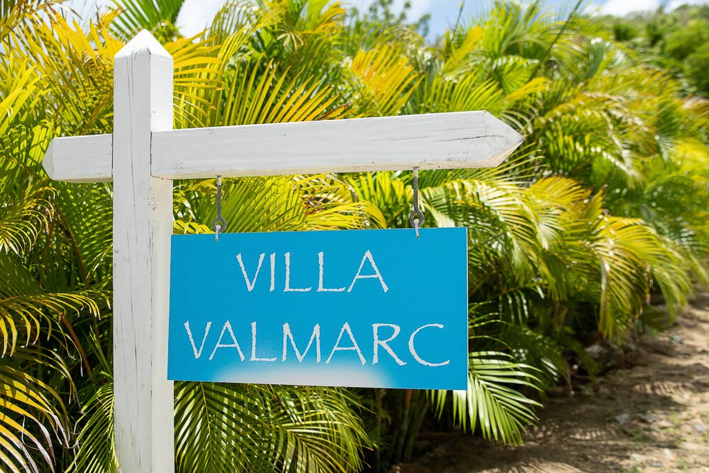 White sign post with a blue sign with Villa ValMarc on it against a backdrop of bright green ferns on a sunny day.