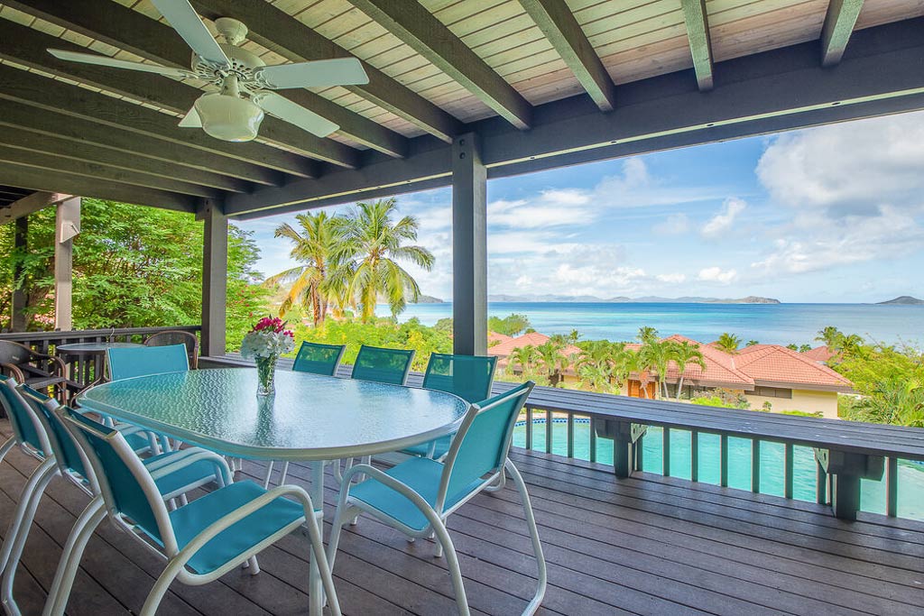 Outdoor dining area on a covered deck with a ceiling fan at Villa Del Sole with Mahoe Bay in the background on a sunny day.