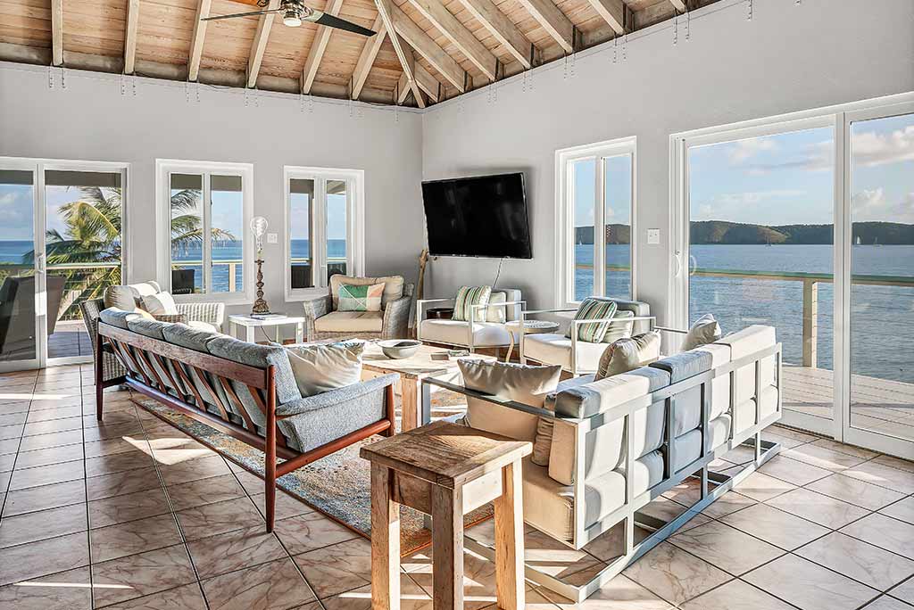 Spacious living room with chairs, couches and a flat-screen TV with large windows looking out on the bay in the background.