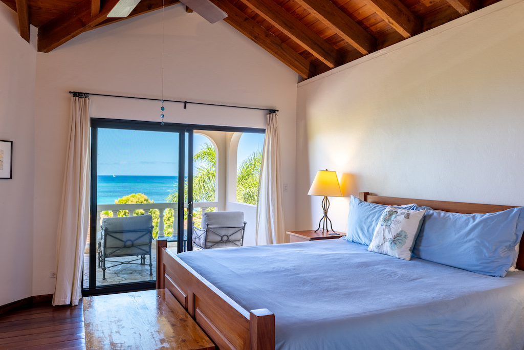 Guest room with a king bed and side table with a lamp next to a glass door to a patio with Mahoe Bay in the background.