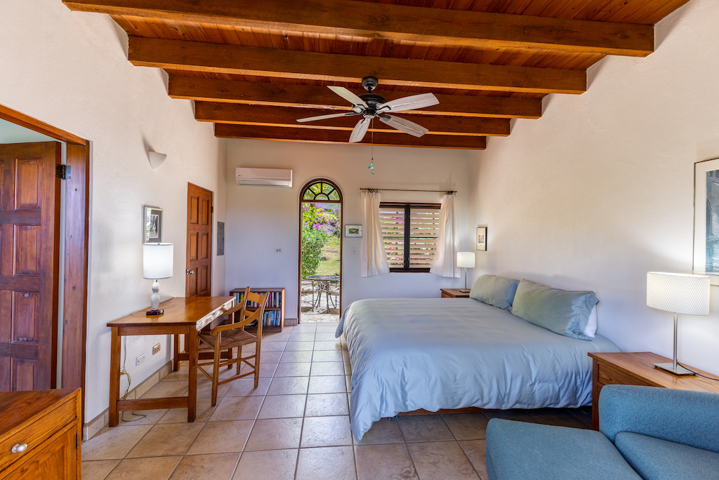 Guest room at Bellamare Villa with a king bed, desk with chair, tile floor, a wood-beam ceiling and open door to a patio.