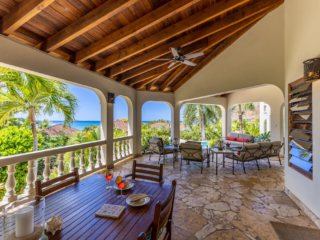 Bellamare Villa: Covered Patio Covered natural-stone patio at Bellamare Villa with a dining table and living area and a backdrop of palm trees and the sea.