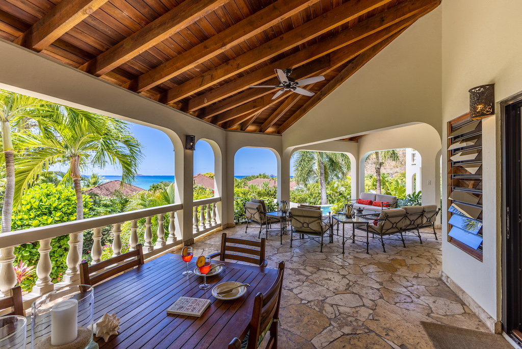 Covered natural-stone patio at Bellamare Villa with a dining table and living area and a backdrop of palm trees and the sea.