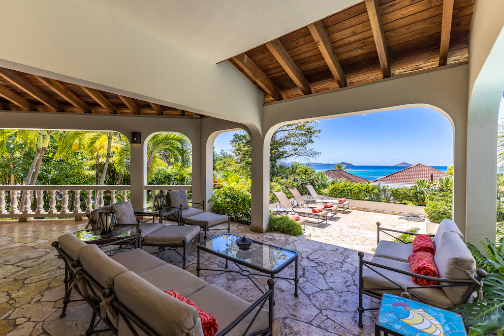 Covered stone patio with outdoor living area at Bellamare Villa with a sun deck and the water of Mahoe Bay in the background.