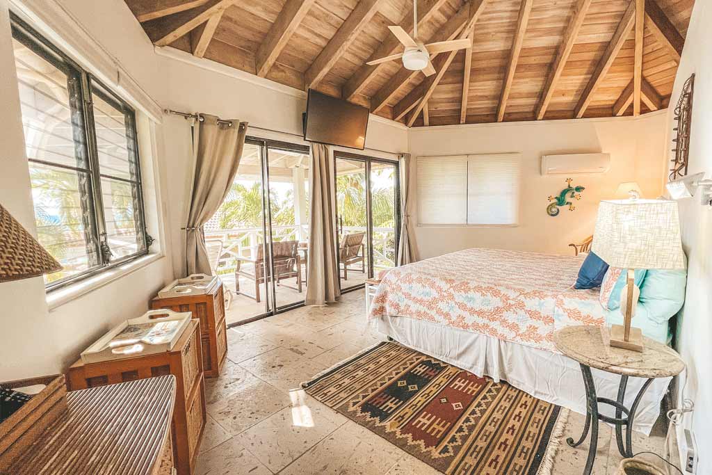Sea Fans Villa master bedroom with a king bed, wood-beam ceiling with a fan, mounted TV and glass doors to a covered patio.