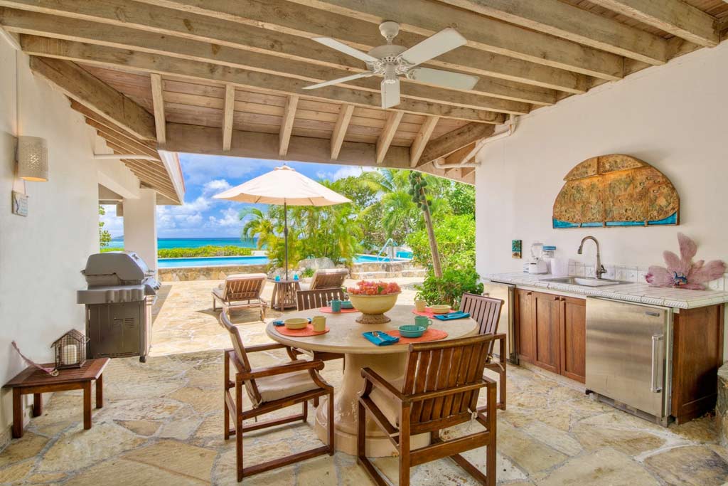 Outdoor dining area with a round table and chairs, a wet bar and BBQ grill and a pool and blue ocean water in the background.