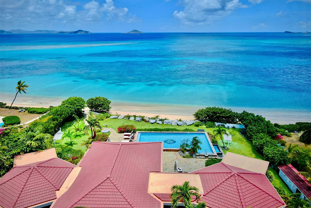 Red tile roof top of Sea Fans Villa with a pool and grounds beyond and a beach and pristine Mahoe Bay in the background.