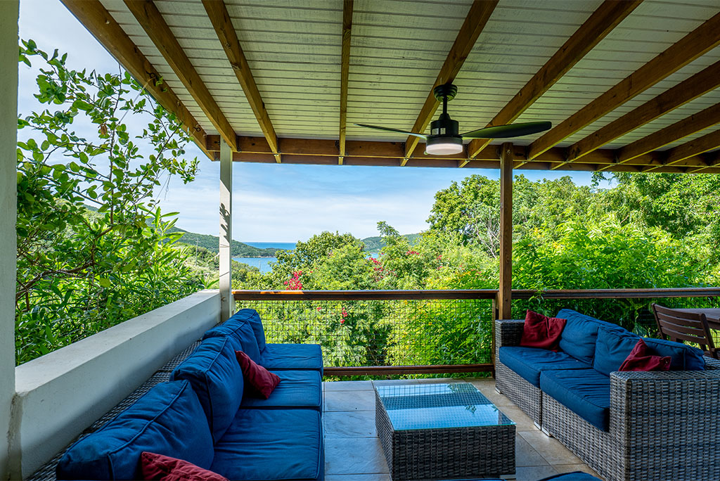 Tamarind Villa’s covered patio with wicker couches with blue cushions and green treetops and Leverick Bay in the background.