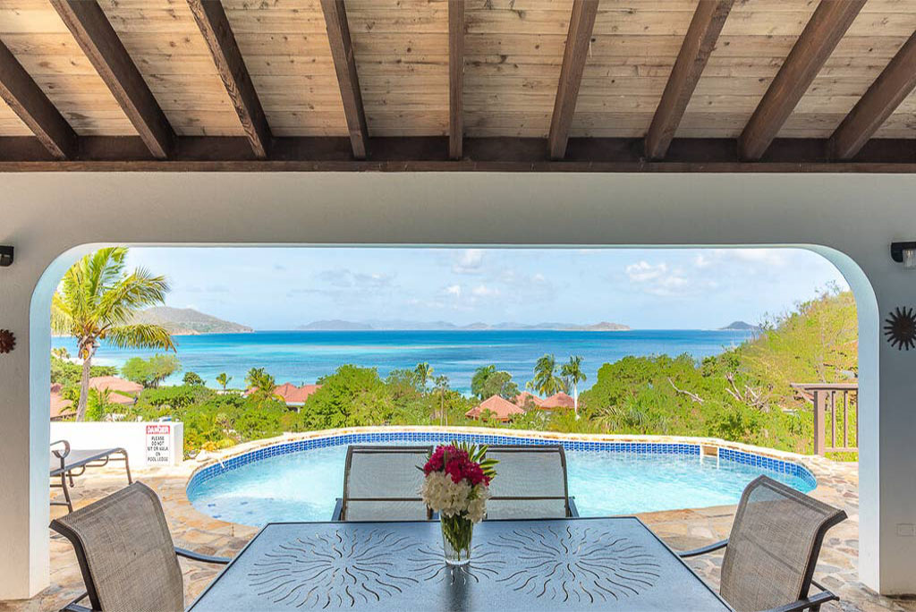 Table and chairs on stone patio with a pool and Mahoe Bay in the background.