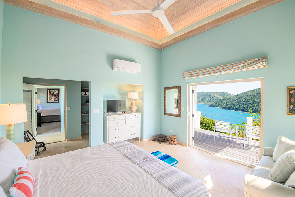 Guest room with a king bed, aqua-blue walls, wood-bean ceilings and open door to a patio with Leverick Bay in the background.