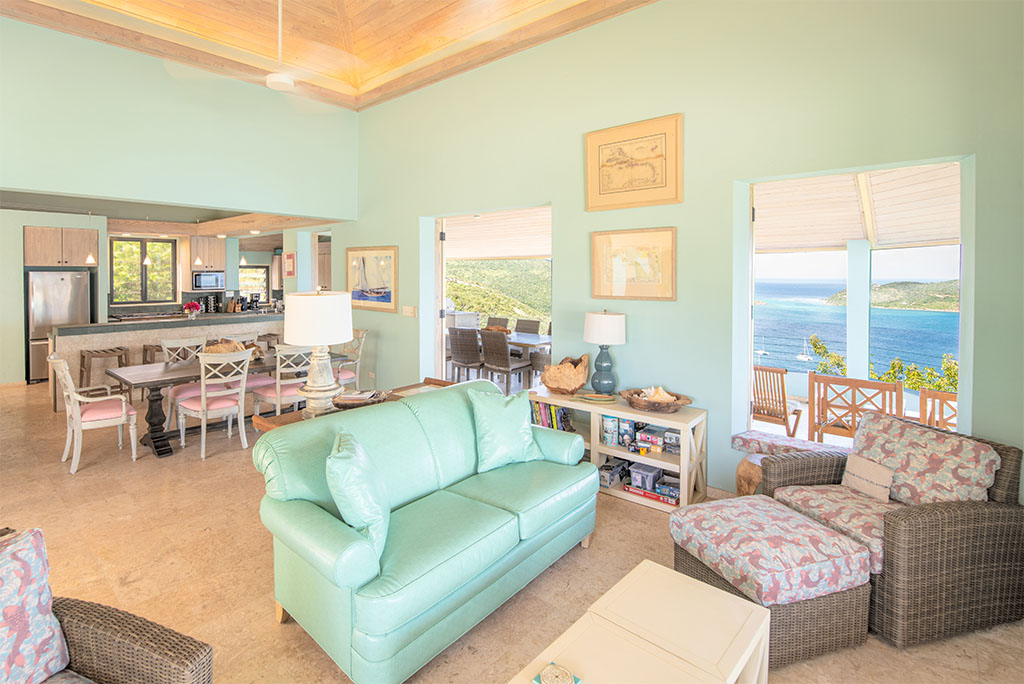 Sitting area with a cushioned loveseat and wicker chair with an open kitchen in the background and views of Leverick Bay.