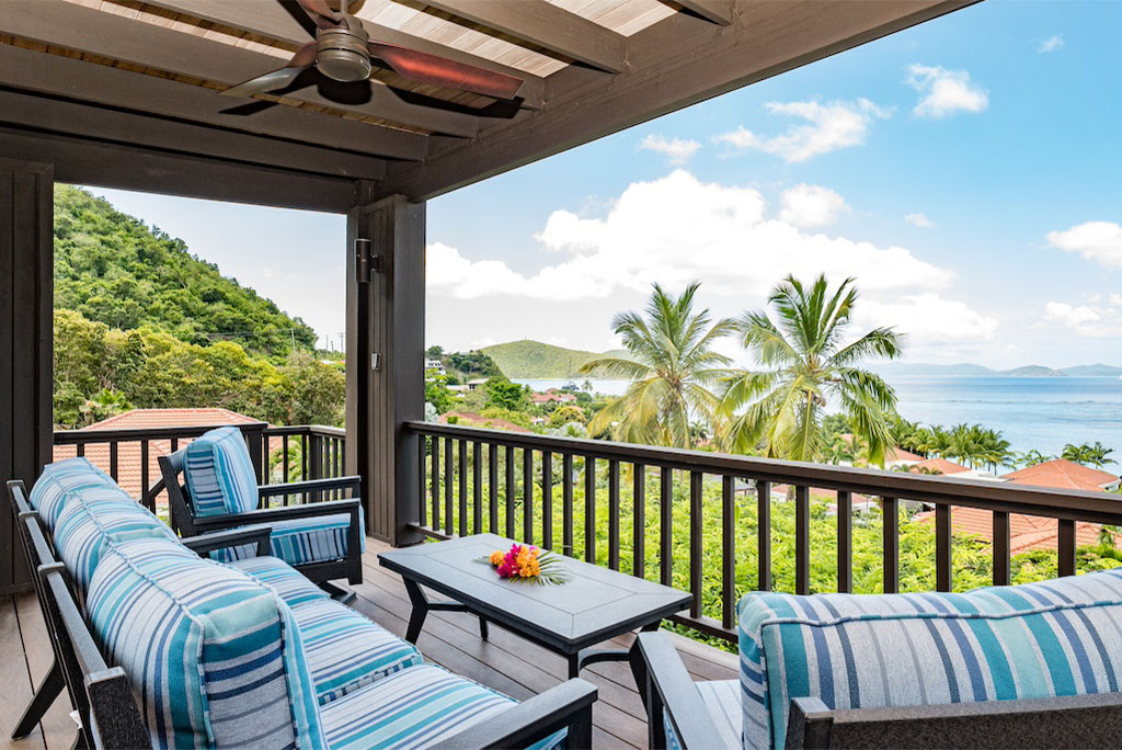 Viewing deck with colorful patio furniture and a wood-beam ceiling with a fan and palm trees and Mahoe Bay in the background.