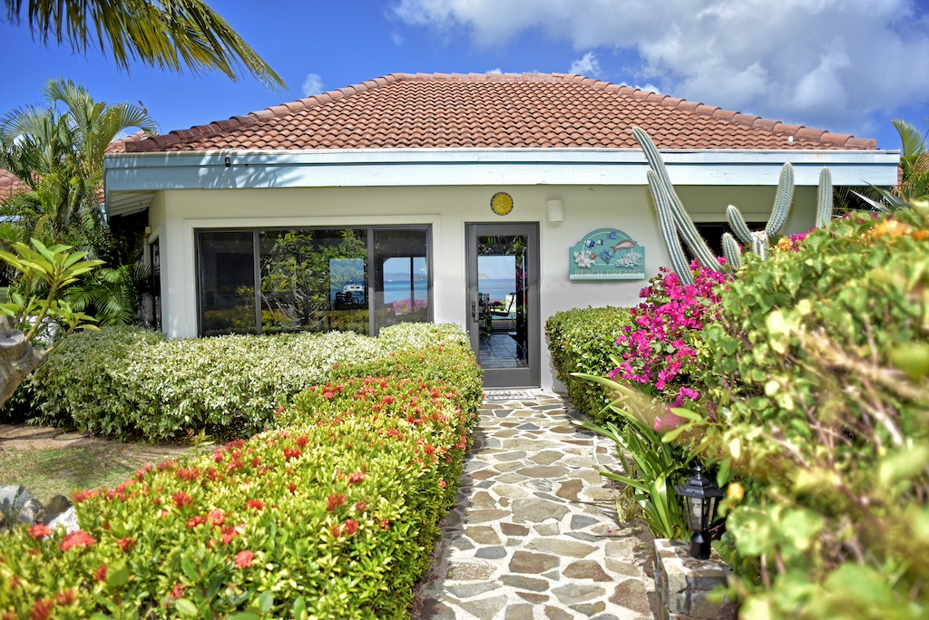 The approach to Beach Dreams villa with a stone path bordered by tropical hedges leading to the front door and blue skies in the background.