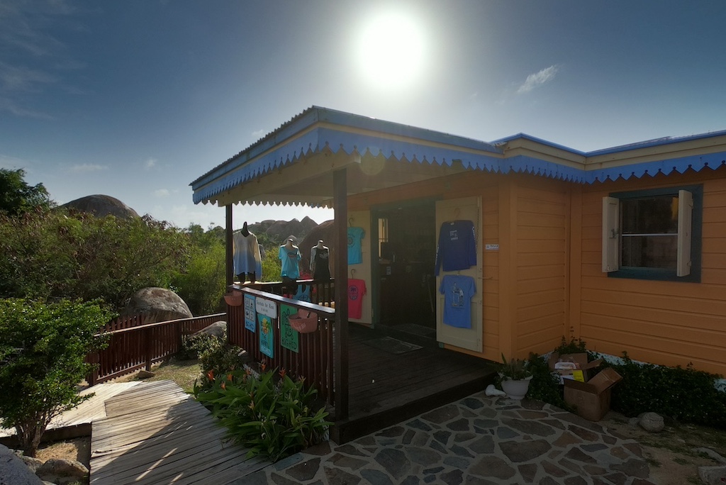 A small shop with yellow walls and blue trim displaying shirts for sale on the front porch with the bright island sun in the sky above.