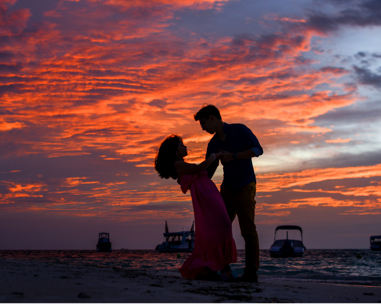 A couple dance hand-in-hand on the beach at sunset against a darkening sky painted by pink clouds. 