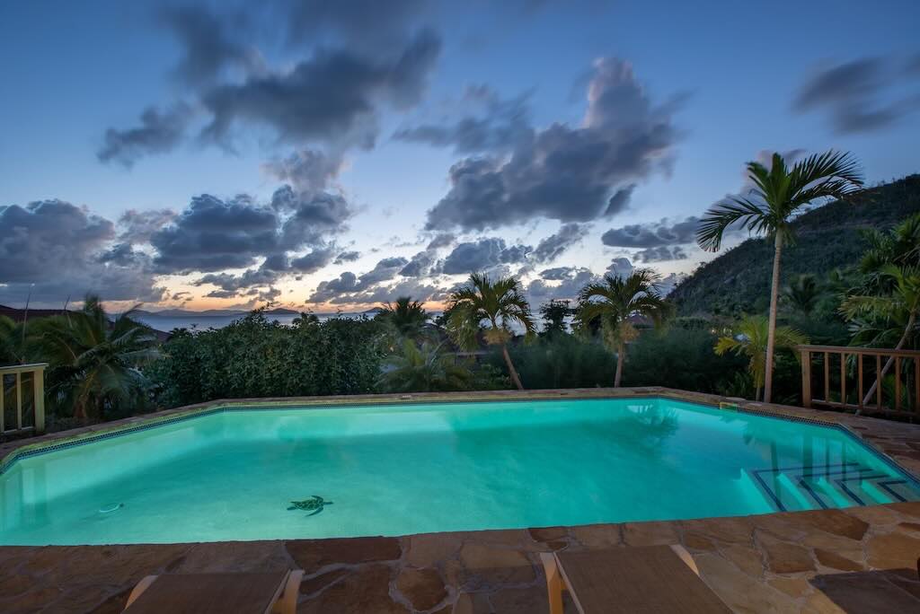 Loblolly Pool Night Large swimming pool surrounded by a stone patio with a backdrop of palm trees and a party cloudy sky at sunset.