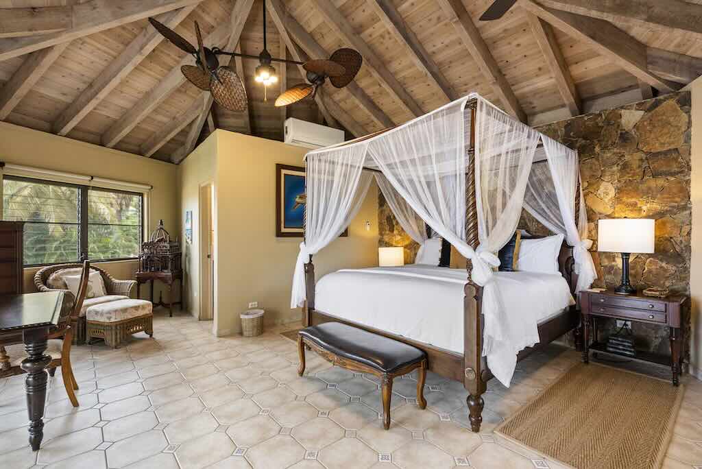 Sandcastle Villa_Guest Suite Private bedroom with a king-sized four-post bed sitting on light tile floors and a stone wall in the background with a wood beam ceiling with fans.