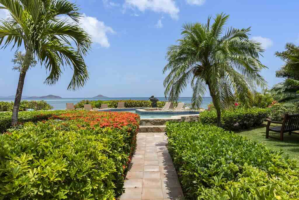 Sandcastle Villa_Pool Garden A stone tiled path cuts between green hedges to a swimming pool surrounded by lounge chairs with Mahoe Bay in the background.